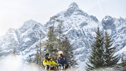 Couple sitting on sleds in snow with snowy mountains and pine trees behind