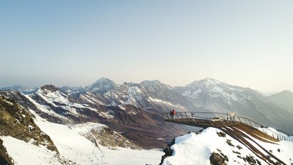 Aussichtsplattform mit zwei Menschen auf schneebedecktem Berg mit Alpen im Hintergrund